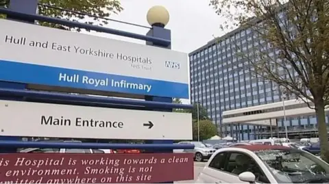 A blue and white sign directing visitors to the main entrance of Hull Royal Infirmary. The tall, glass-fronted hospital building is in the background. Cars are parked nearby.