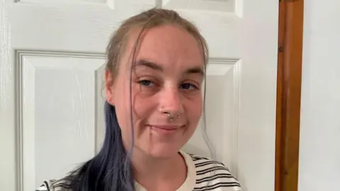 A young woman with dark long hair pulled back in a ponytail smiles into the camera. She is standing in front of a white paneled door and is wearing a top with narrow white and black stripes and has a nose ring.
