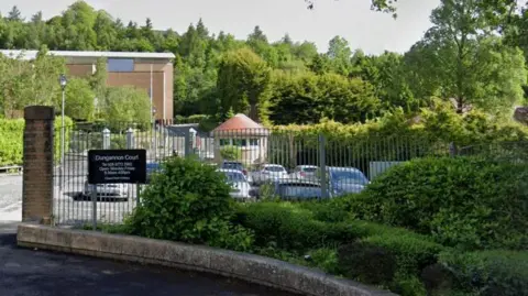 A car park with a grey fence and bushes in front of it. There is a black sign on the fence reading Dungannon Court. There is a beige building and trees behind the car park.