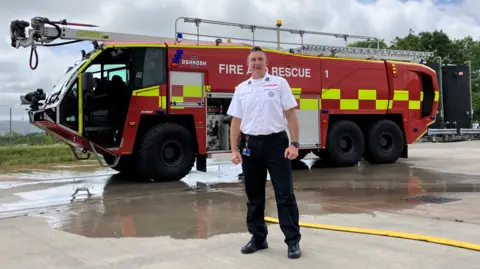 A fire officer stands in front of a large fire engine. He is wearing a white shirt and black trousers. The vehicle is red with yellow markings. On its roof is a ladder and a extendable arm for targeting fires with water or foam. 