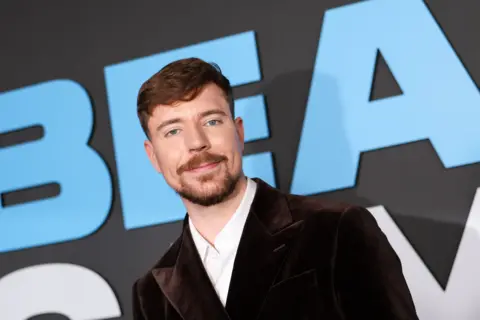MrBeast aka Jimmy Donaldson has light brown hair, a beard and moustache. He is wearing a dark-coloured velvet suit jacket.