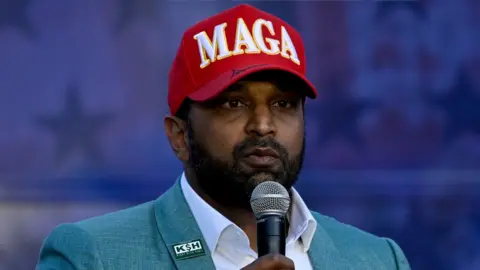 Kash Patel holds a microphone. He is wearing a green blazer and a red hat that says "MAGA" across the top. It appears to have Donald Trump's signature on the brim