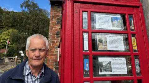A man is standing next to a old red phone box. He is wearing a blue check shirt and a navy jacket. On the phone box there are a series of white notices with photos, showing places of interest. 