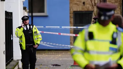 PA Media A uniformed police officer uses a mobile phone while walking past a cordon on part of the street. Several out-of-focus police officers stand in uniform on the right of the image