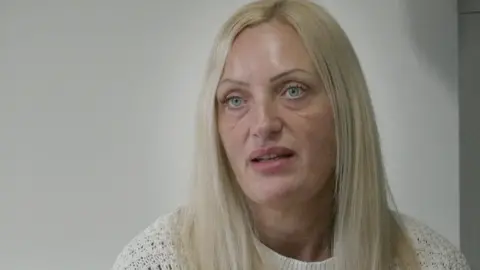 Jenni Smith with long blond hair wearing a white knitted crew neck jumper. She is sitting in an office with a white wall behind her. She is looking impassive.