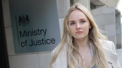 Martis Media Harper has long blonde hair and blue eyes. She wears a white coat and stands next to the sign for the Ministry of Justice.