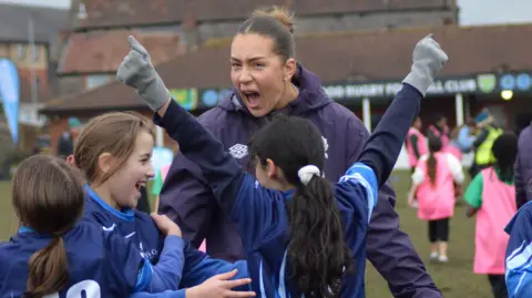 An adult female rugby player in a blue kit cheers with her mouth wide open as she celebrates with younger players, also in blue, as part of a training session