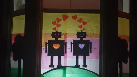 Window decorated with a colourful tissue paper design of two robots with hearts between them