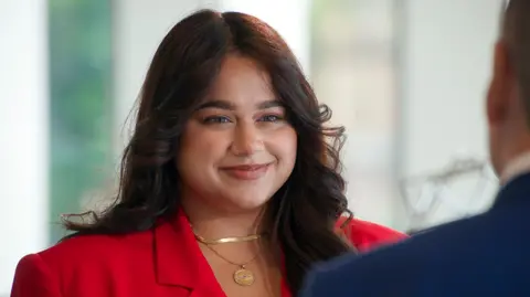 PA Media Close‑up of Karishma Vijay wearing a red blazer during a filmed interview. She is facing someone off camera in a business setting, with light coming through windows in the background.