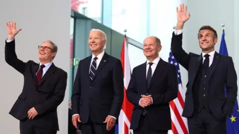 Reuters UK Prime Minister Keir Starmer, German Chancellor Olaf Scholz, US President Joe Biden and French President Emmanuel Macron pose for a family photo session at the Chancellery in Berlin, 18 October. Starmer and Macron are holding their right hands in the air and waving. All four are smiling and wearing dark suits, and there are flags of the UK, US and EU behind them.