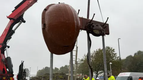 An orange-brown coloured helmet made from steel hangs from a hook suspended by silver steel poles.