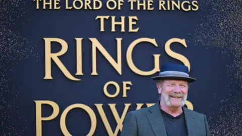 Getty Images Peter Mullan poses on the red carpet upon arrival to attend the world premiere for season two of "The Lord of the Rings: The Rings of Power" at the BFI Southbank, in London. He stands against a huge backdrop with the show's title on it and wears a dark grey suit jacket, black t-shirt and a straw hat in navy with a coloured band.