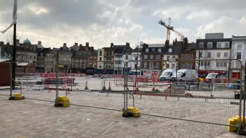 Pete Cooper/ BBC A wide shot of the market square undergoing construction with cobbled flooring, metal gates, cones, vans and cranes on show