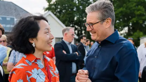 Yuan Yang Yuan Yang talking to Sir Keir Starmer during the 2024 general election campaign. She has medium length dark hair and is wearing a red flowery shirt, whilst he is wearing a blue shirt, glasses and has grey slicked over hair.