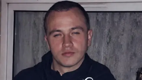 A man with very short dark brown hair is wearing a dark coloured coat while part of a trophy he is holding in his hand can just be seen. He is standing in front of a window.