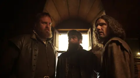 BBC Studios Three actors portraying Guy Fawkes, Thomas Wintour, and Robert Catesby. They are dressed in historical dark clothing, in a room, with a window, which is dark. They all look tense and serious. 