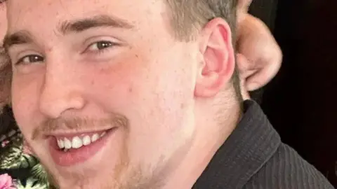 A close-up photo of a smiling young man, with light brown hair and a thin beard. 