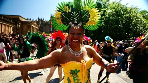 BBC A carnival performer wearing a large green and yellow feathered headdress and a yellow-and-green costume walks at the front of a colourful street parade. Crowds and other performers fill the background on a sunny day.