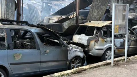 In the foreground, there are two severely fire-damaged vehicles parked beside a kerb: A grey MPV on the left has extensive fire damage, with burned paintwork, missing windows and a heavily charred roof.