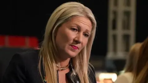 BBC Laura Anne Jones wearing a black top, in conversation with a person slightly off camera. She has a black backdrop behind her and is wearing a Reform party lanyard.