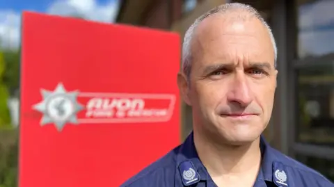 Steve Quinton looking at the camera while wearing his Avon Fire and Rescue uniform which is blue with white patches. Behind him, but not in focus, is the service's logo on a large red sign