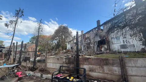 A row of residential properties is shown after a major fire, with extensive damage visible across several homes. Charred walls, burnt-out rooms and blackened rooflines can be seen, while the foreground contains scorched fencing, debris and the remains of trees or garden structures that have been reduced to blackened posts. A ladder leans against one of the damaged buildings, and scattered household items remain among the wreckage. The scene is photographed in daylight under a bright blue sky, highlighting the scale of the destruction.