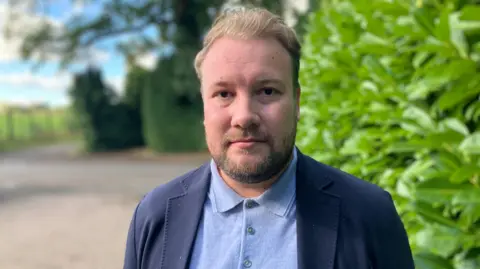 A man wearing a light blue polo top and blue blazer, with bushes and trees behind him