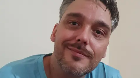Darren King, with greying short hair and a stubble, smiles at the camera. He is wearing a turquoise T-shirt.
