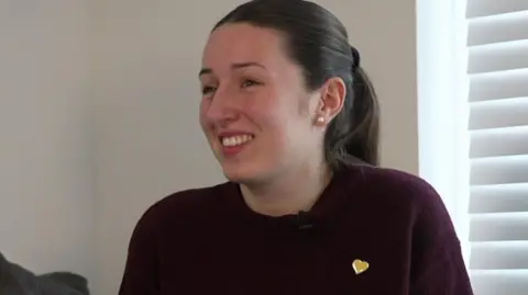 Young woman, 25, wearing a burgundy top with chestnut brown har tied back. She is wearing a yellow heart pin badge on her top.