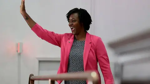Angie Nixon in black and white dress and pink blazer waves her right hand while smiling