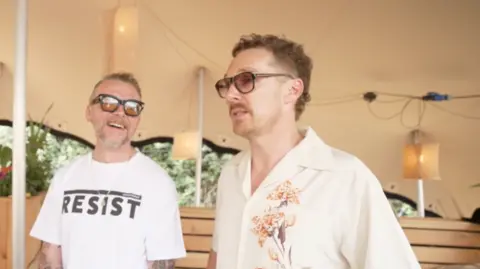Simon Pegg and Benedict Cumberbatch stand side by side under a tent canopy. Pegg is smiling and laughing as he looks towards Cumberbatch. They are both wearing sunglasses. Pegg has on a white T-shirt which says 'Resist' in upper case, black letters. Cumberbatch has on a cream, short-sleeved shirt with a floral design on one side.