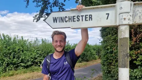 Miles Eades Miles Eades holding a sign which says Winchester 7 miles away. He's wearing a sports top and is on a country lane
