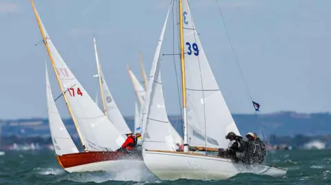 Cowes Week Paul Wyeth Two sailing boats race through choppy water, with spray rising from the waves as crew members sit on the edge of the hulls.