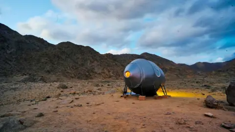 AAKA Space Studio A circular pod is seen parked in the mountains of Ladakh, it is sliver in colour and held up with little pole structures held in place with plywood boxes, with the browny-red backdrop of the Ladakh mountains in the background and some clouds in a dark sky.