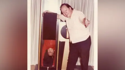 Supplied A magician stands beside a large box that appears to divide someone in half. The lower section of the box shows a person's head and shoulders.
