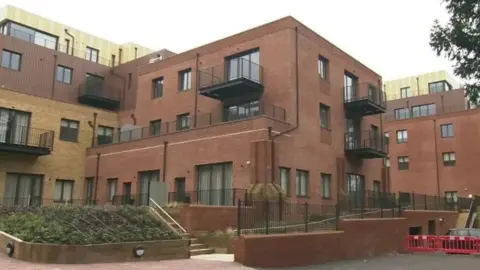 A large brick block of flats, some of which have balconies. The building is three stories and has stairs and a ramp leading up to it.