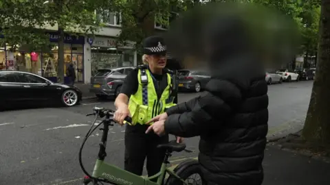 BBC A female police officer called Sgt Clare Moore holds an illegal e-bike on a Plymouth street during a police operation. She is talking to a person - possibly the bike's rider - who is wearing a black coat with its hood covering his head. His head has been blurred. Sgt Moore has a yellow high-vis police jacket on. The e-bike is green and has emovement written on it in white letters. Its wheels and handlebars are black.