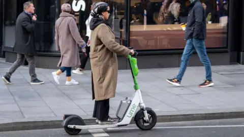A woman rides a Lime electric scooter on a city street in front of a Starbucks, while pedestrians walk past and customers sit inside the café.
