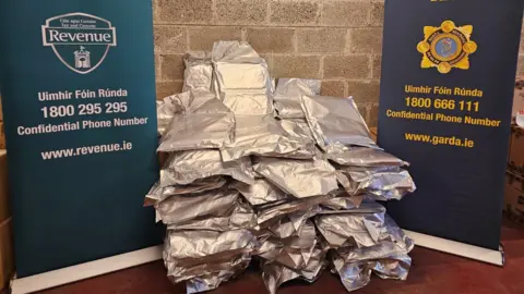 Two large posters are next to a large seizure of drugs which is in silver packets. The posters are dark blue and turquoise with 'revenue customs service' and 'An Garda Síochána' written on it with their logos.