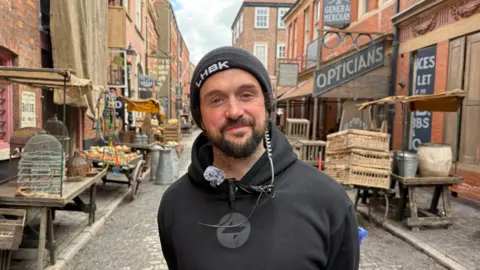 BBC News A man in a beanie hat smiles as he stands in a cobbled street lined with Victorian warehouses and former shops and houses, which has been made up to look like Victorian London, with old signs and period street furniture. The man is wearing a black hooded jumper. He has a black beard and brown eyes. He is also wearing a wire connected to a walkie talkie to communicate with colleagues. 