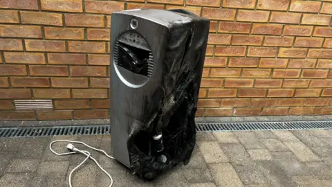 A black and burnt-out air conditioning unit on a driveway.