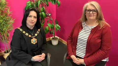 Councillor Eileen Callear, Telford and Wrekin Mayor and Jane Lees, town clerk for Hadley and Leegomery Parish Council sitting together in front of a pink wall with green plants in the background. Both looking into the camera. 