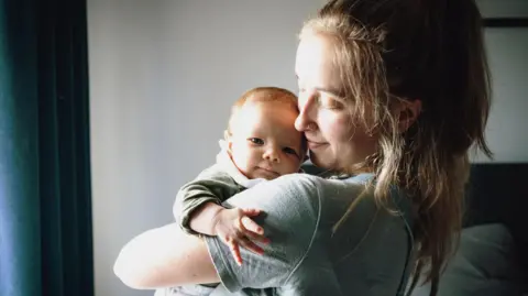A young mum with light brown hair and wearing a grey T-shirt holds her baby in her arms and looks at him with her head turned to one side, standing in front of a window with daylight streaming through.