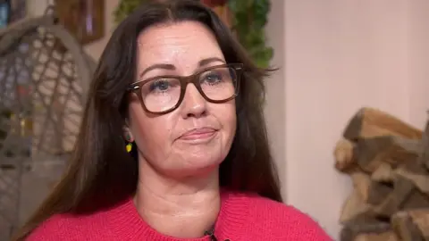 Mark Norman BBC A woman with long dark hair wearing glasses and a red jumper in a living room.