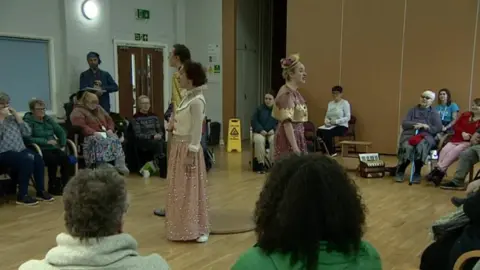 Three people standing up in the middle entertaining the people at the centre sitting around them. Two of them in the middle are wearing long dresses.