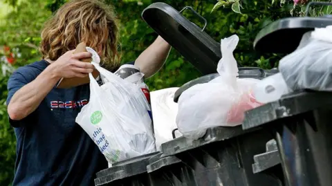 A man with long hair, his face obscured, empties waste into a wheelie bin 