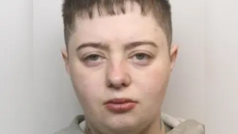 A mugshot of a pale person with short hair, which is buzzed at the side and slightly on the forehead from above. Her eyes are slightly narrowed and she has a neutral or tired expression. She has a tattoo on the side of her neck and a grey hoodie on.
