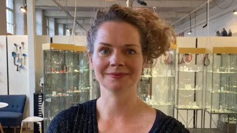 A woman wearing a dark blue cardigan and long hair gathered to one side of her head standing in a jewellery gallery with glass display cabinets.