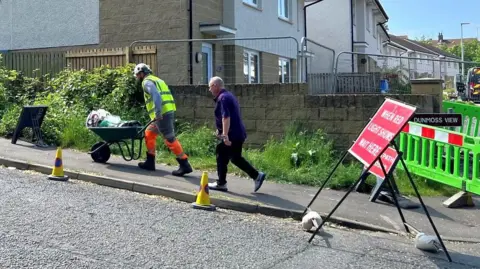 A man in hi vis with a wheelbarrow with household items followed by another man
