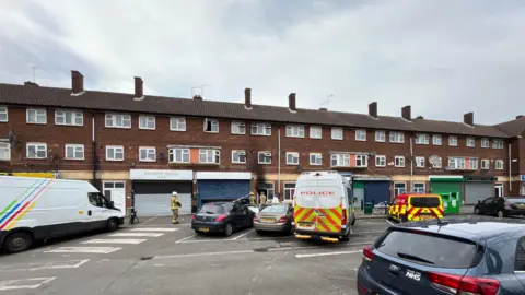 Multiple vehicles parked outside a terraced building of flats and shops. A police van and fire service vehicle are two of the vehicles parked outside. Firefighters can be seen by the entrance of one of the flats, which has black scorch marks over the door.